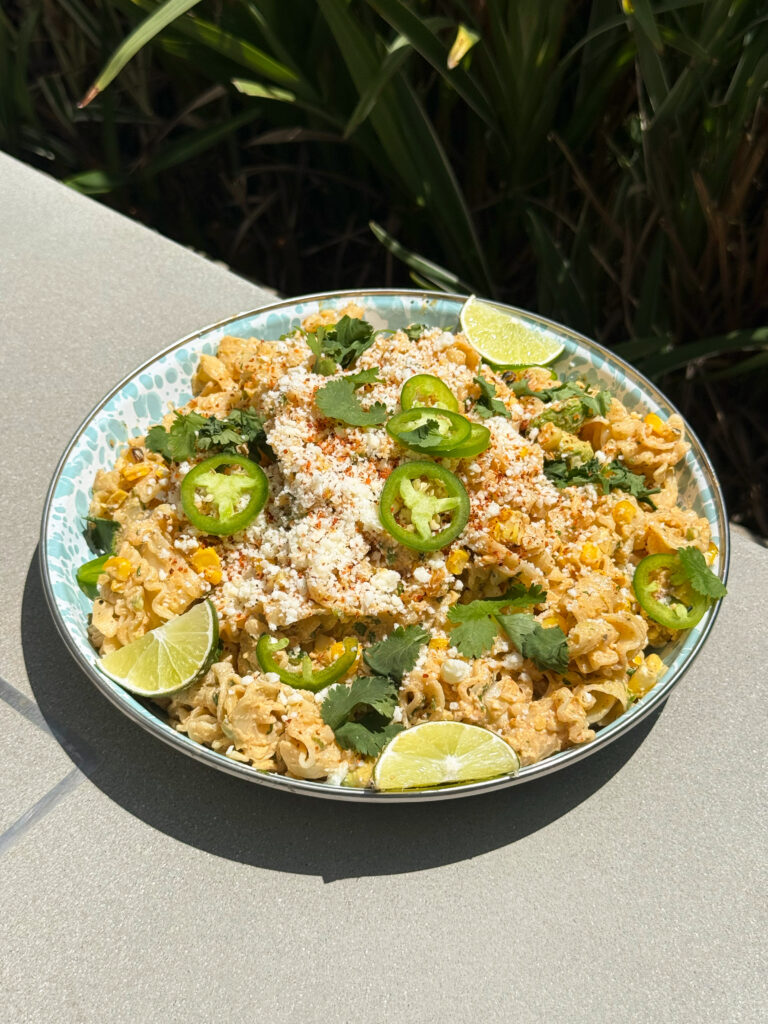 Elote Pasta Salad sitting on the edge of a table with greenery in back